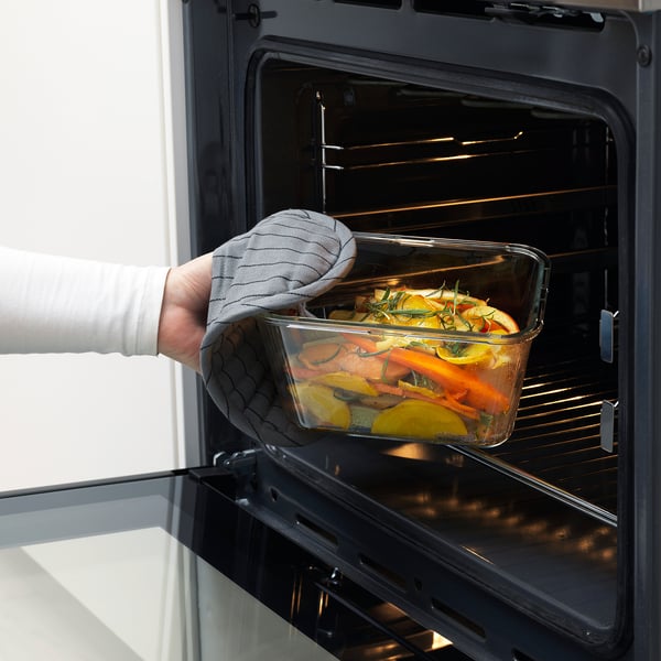 Person inserting glass food container, filled with vegetables, into an open oven, using an oven mitt.