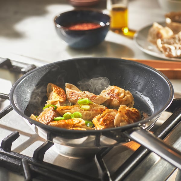 Stir-fry pan on a stovetop cooking dumplings. Steam rises, showing its hot. Green onions garnish the food.