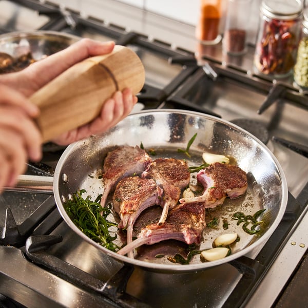 Cooking lamb chops in IKEA 365+ pan on gas hob. Wooden handle holds pan steady. Garlic and herbs sizzle with meat.