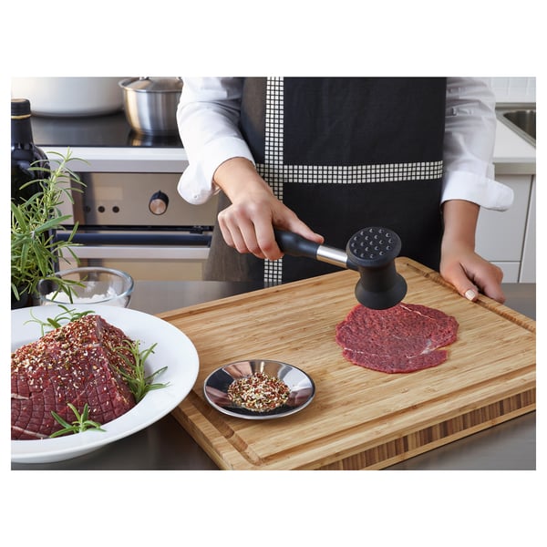 Person uses meat hammer on cutting board with meat. Small bowl of seasoning nearby. Kitchen setting.