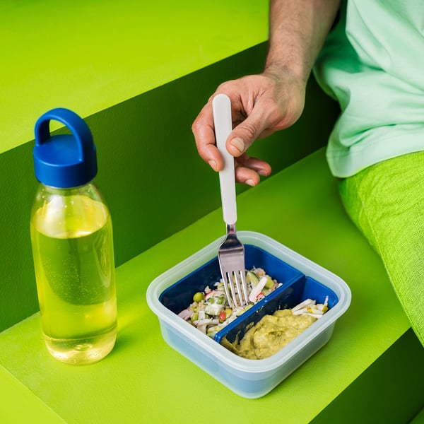 Person eating from a blue compartment lunchbox with a fork, next to a clear yellow liquid IKEA 365+ bottle with a blue lid on a green surface.