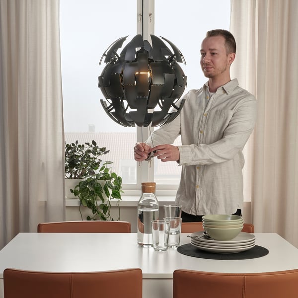 Person adjusting strings of a black globe-shaped pendant lamp above a dining table set with plates, glasses, and a water jug.