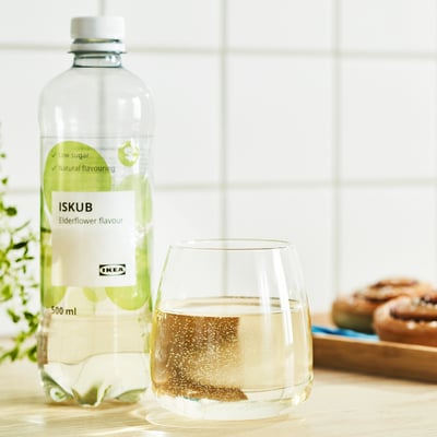 A clear bottle of IKEAs ISKUB elderflower flavoured drink stands next to a filled glass on a table, surrounded by pastries and greenery.