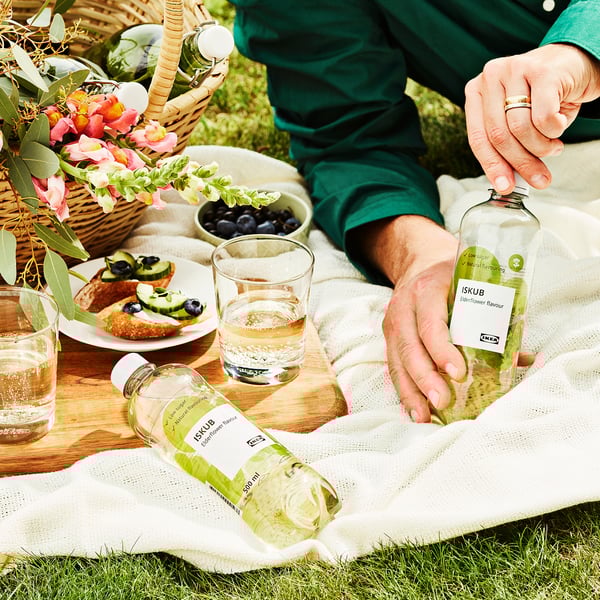 Person pours ISKUB soda outdoors. Basket with flowers and snacks beside them.