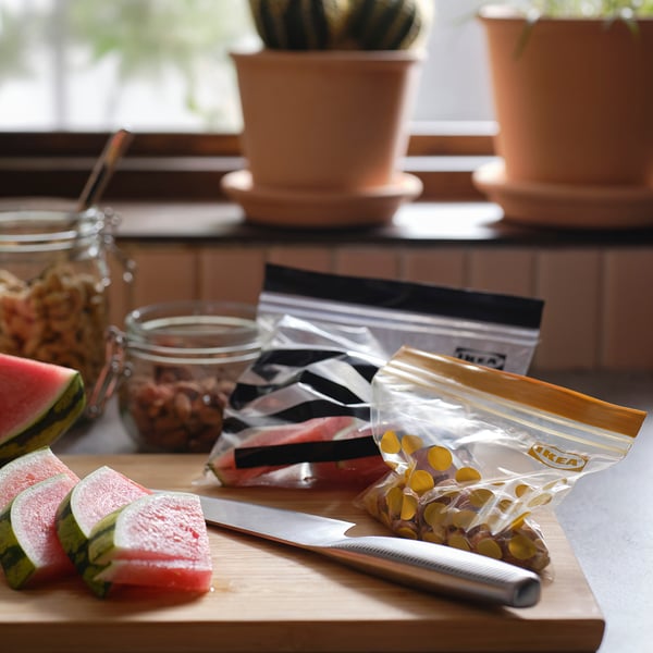 Wooden cutting board with watermelon slices, knife, sealed food bags, jars, and potted plants.