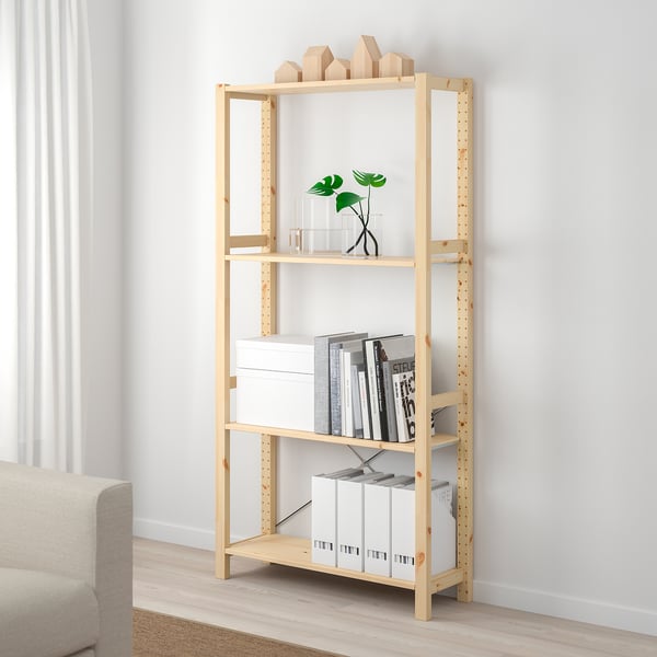 An IVAR light brown pine bookshelf. It stands tall next to a beige sofa, holding books, boxes, and a plant.