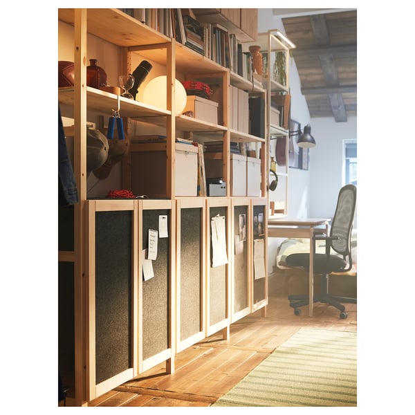 Home office with IVAR wooden storage shelves. Light wood shelves hold books, boxes, and vases. Felt-covered doors with pinned notes add warmth.