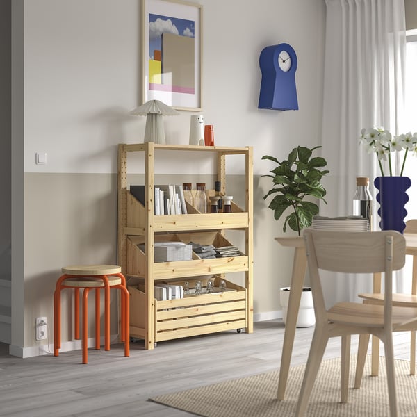 Modern living room with minimalist IVAR pine storage shelf against wall, wooden chair, vibrant orange stool, blue clock, plants, books, and vases.