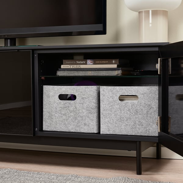 Black tv stand with open doors, grey felt storage boxes, books, and a lamp.