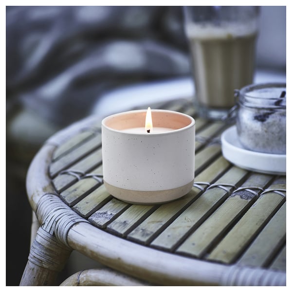 Ceramic candle in white with speckles on wicker table, lit with salt jar beside.