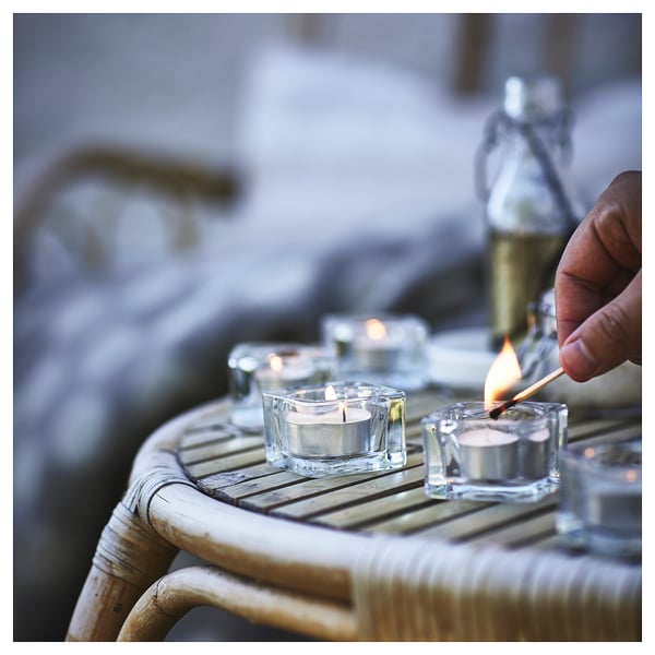 Person lights glass candle holder on wicker table, with other lit candles and a bottle in the background.