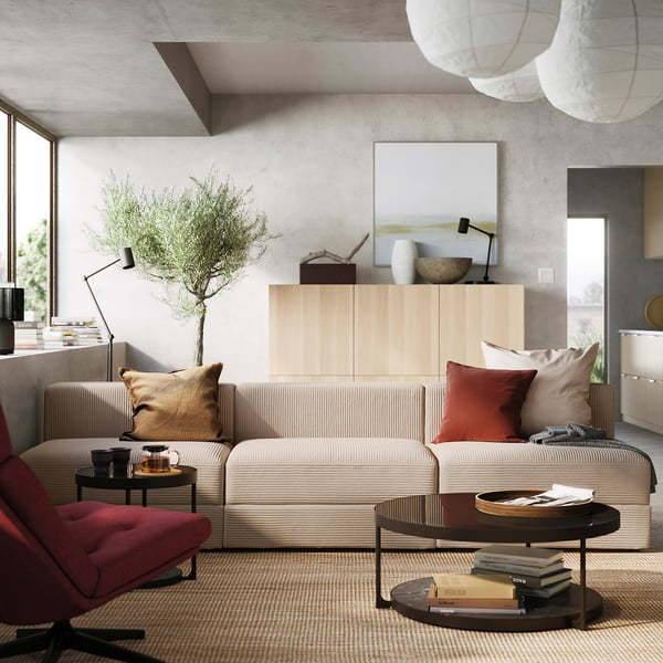 Modern living room with JÄTTEBO modular sofa in beige, wooden cabinet, colourful pillows, plant, and coffee table with books.