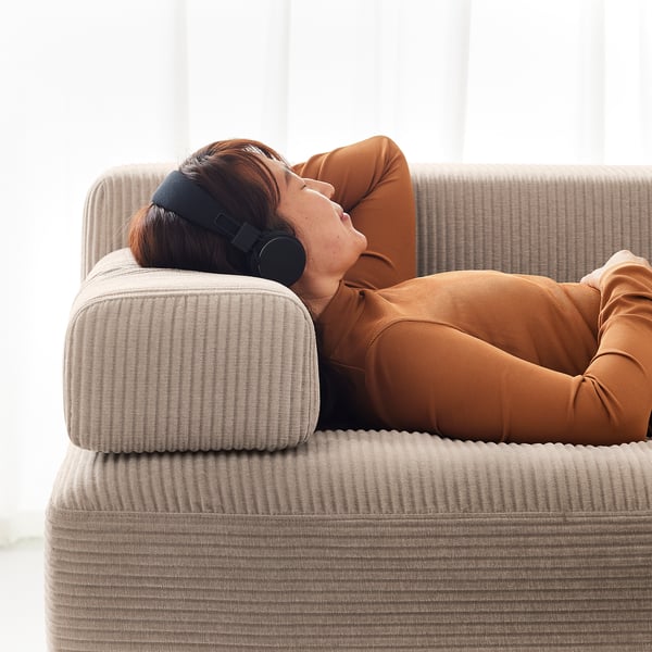 A person naps on a beige JÄTTEBO sofa, wearing a headband and brown jumpsuit.