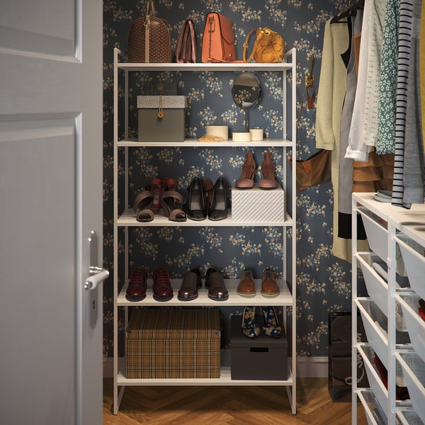 Organised closet with white JONAXEL shelves, holding shoes, handbags, and clothes. Dark floral wallpaper backdrop.