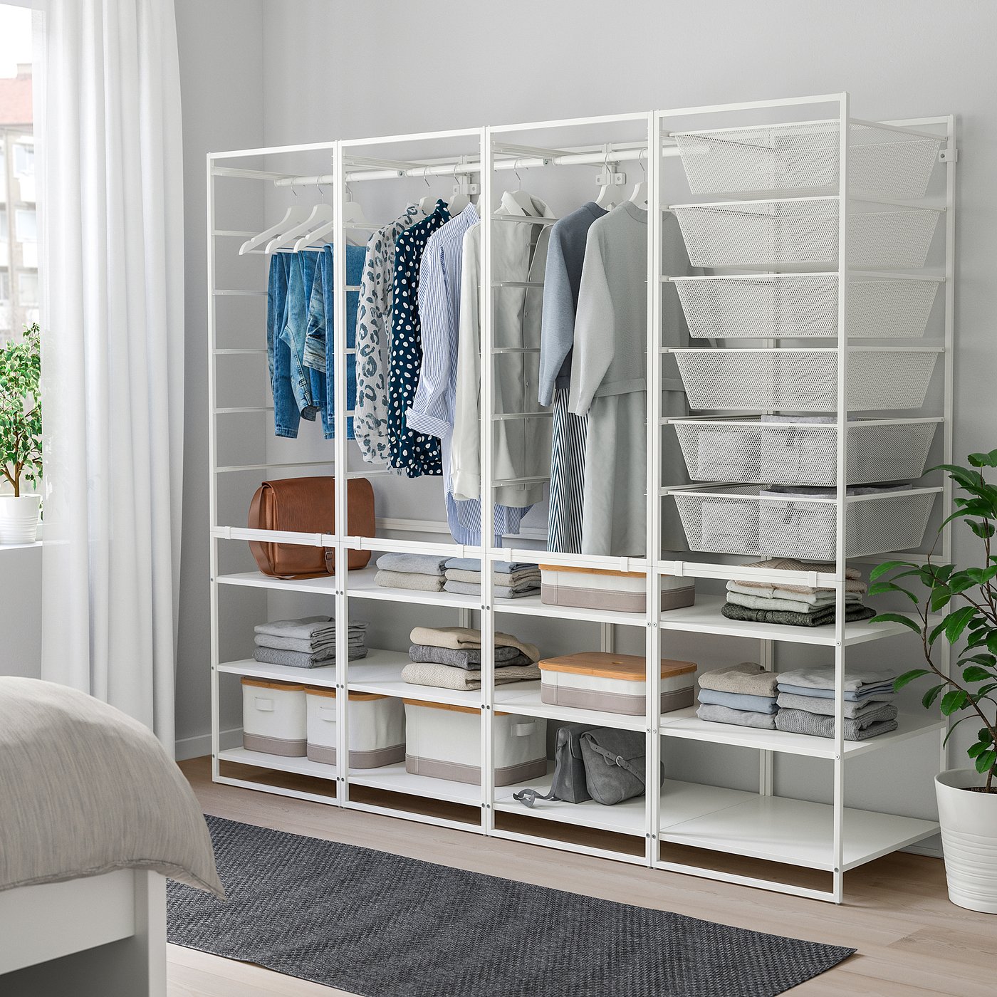 White JONAXEL wardrobe with shelves and rails, filled with clothes, positioned beside bed and plant.