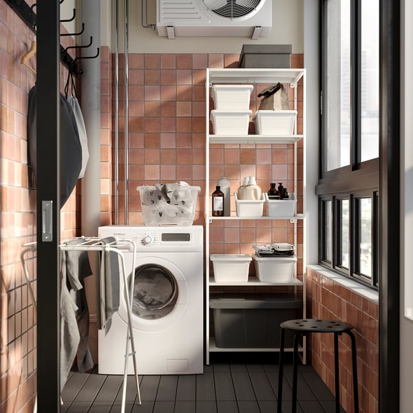 Small laundry room with white washing machine, open shelving unit with cleaning supplies, and tiled walls.