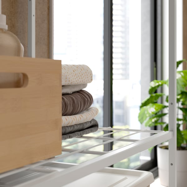 Open shelving unit with neatly folded towels, wood drawer on left, plant on right, near window.