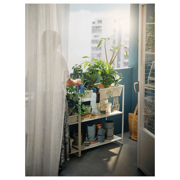 Person watering plants on a white JOSTEIN storage shelf near a window, with curtains and city view.