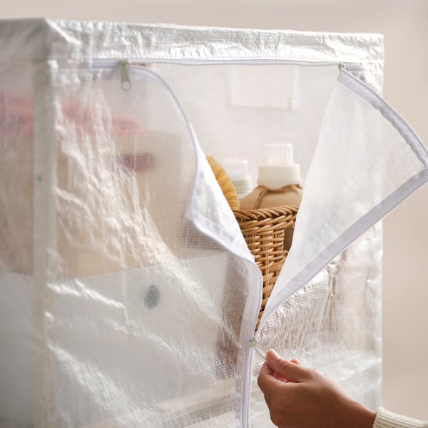 A person zips up a clear JOSTEIN storage bag, containing a basket with bottles.