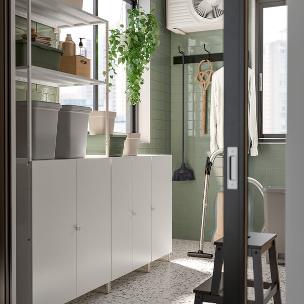 Modern utility room with JOSTEIN white storage cabinets, grey bins, green tiles, hanging plants, and cleaning tools.