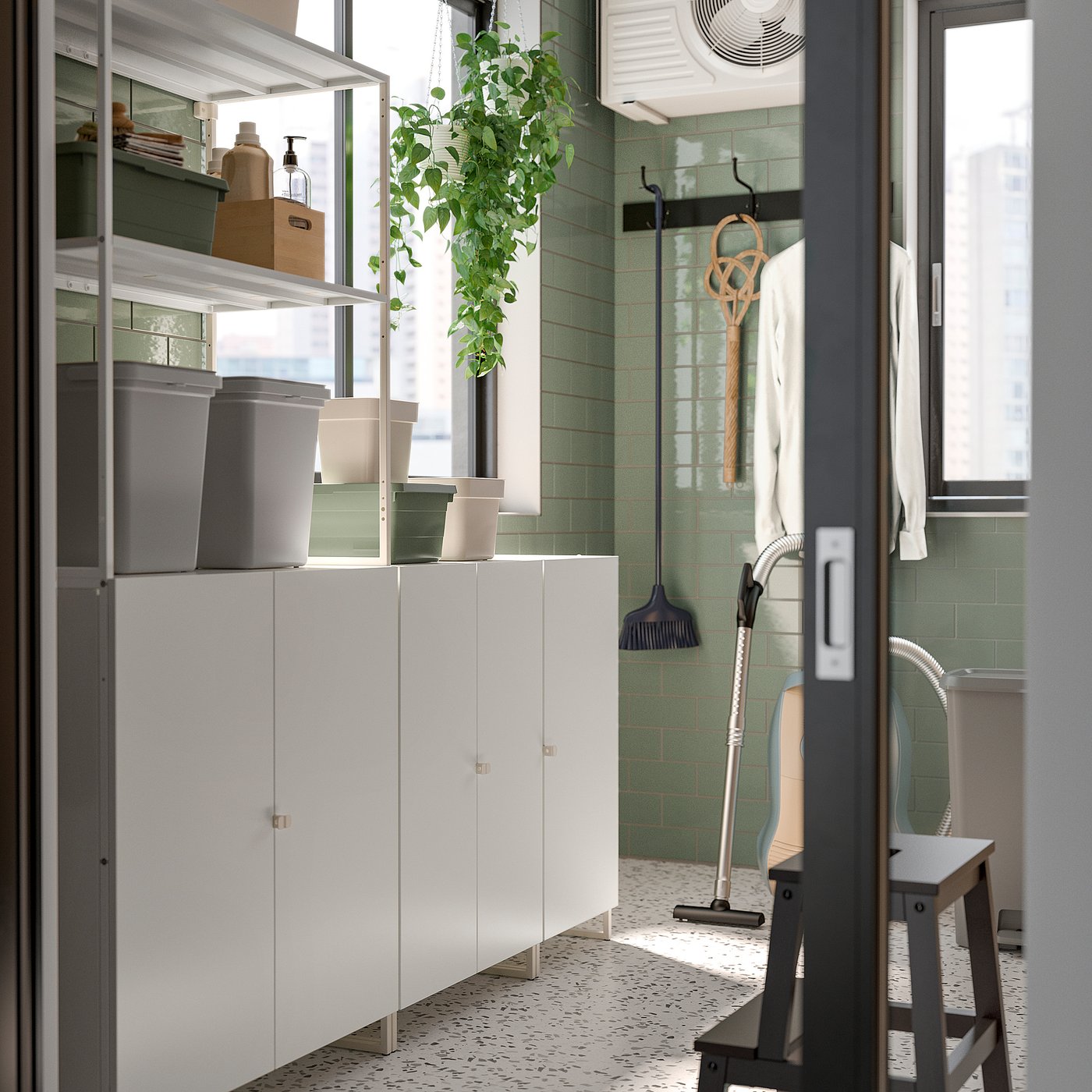 Modern utility room with JOSTEIN white storage cabinets, grey bins, green tiles, hanging plants, and cleaning tools.