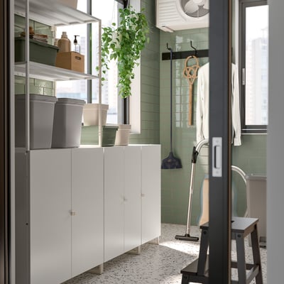 Modern utility room with JOSTEIN white storage cabinets, grey bins, green tiles, hanging plants, and cleaning tools.