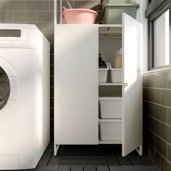 White JOSTEIN storage cabinet in laundry room with black floor, holding plastic bins and cleaning items.