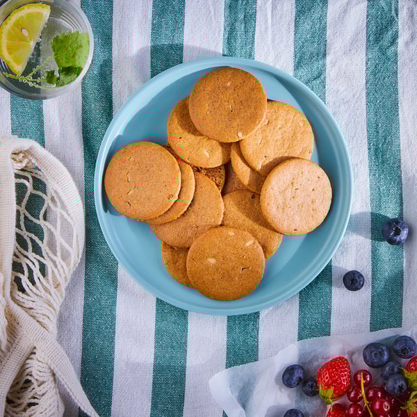 A blue plate of brown ginger cookies sits on a green striped cloth, beside a drink with lemon and mint, and a basket of mixed berries.