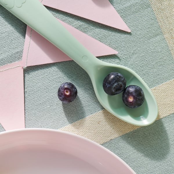Image a pale green kids spoon with three blueberries on it, placed on a geometric-patterned mat with pastel pink and beige triangles.