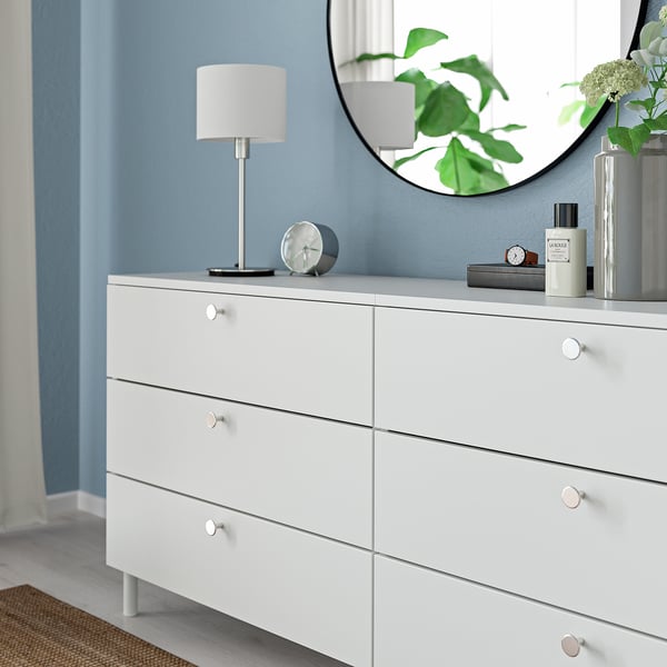 White dresser with drawer knobs, silver lamp, clock, mirror, green plants, blue wall.