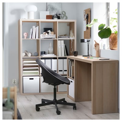 Modern home office with wooden KALLAX desk, black swivel chair, organised shelves, decorative items, and plants.