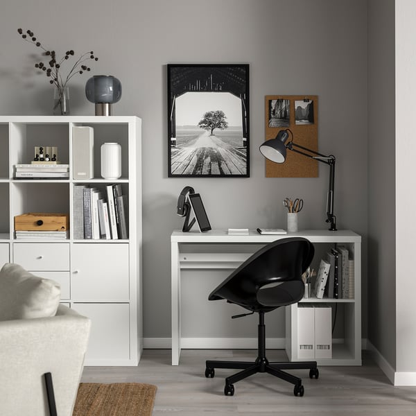 Modern home office with KALLAX desk, black swivel chair, grey wall, bookshelf, photo, lamp, tablet, and sofa.