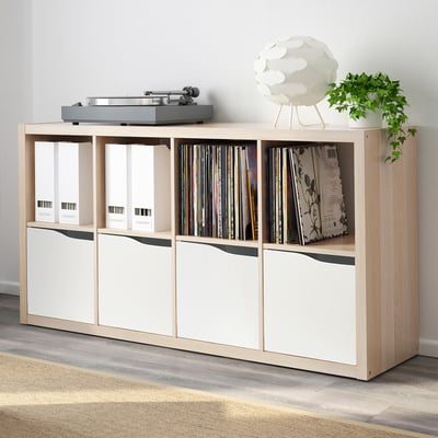 Light-colored KALLAX shelf unit filled with vinyl records and storage boxes. A turntable sits on top, and a white lamp and plant are placed beside it.