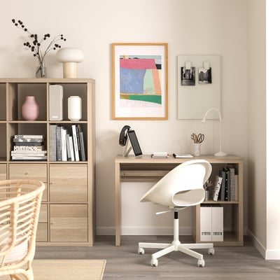 Home office with KALLAX desk, swivel chair, laptop, lamp, artwork, and bookshelf.