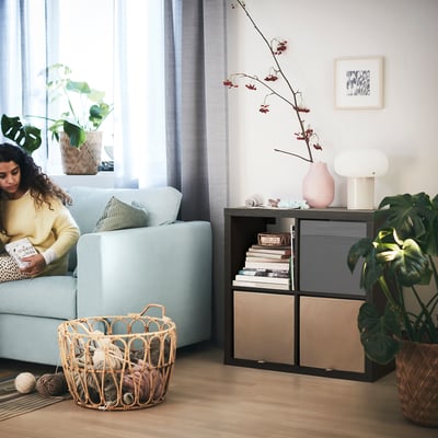 Living room with KALLAX shelving unit and light blue couch. Person reading, yarn basket, and plants.