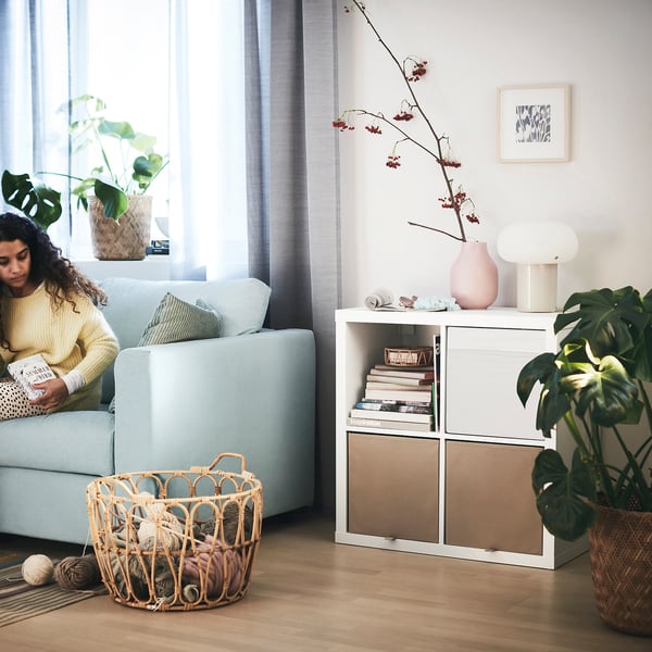 Cosy living room: person reads on blue sofa, KALLAX shelves with books & plants, yarn basket, natural light.