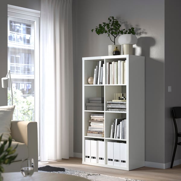 White KALLAX bookshelf with books and decor, by window, in gray living room.