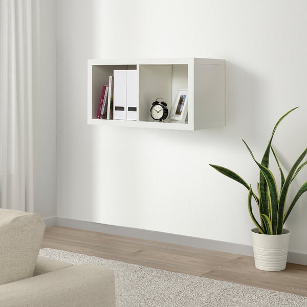 White KALLAX wall shelves hold books, clock, and photo. Clean-lined, minimalist design. Next to a potted plant and chair.