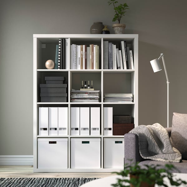 White KALLAX shelving unit with books, files, and decor. Includes a potted plant and floor lamp.