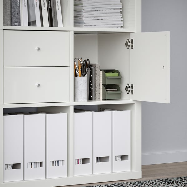 White KALLAX shelving unit with open door revealing stored office supplies; additional shelves hold books and binders.