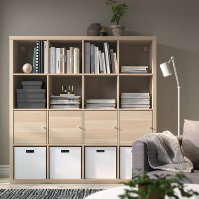 KALLAX bookshelf, light oak, cubic shape, modern design, tall, holds many books and decor. A cosy grey couch rests beside.