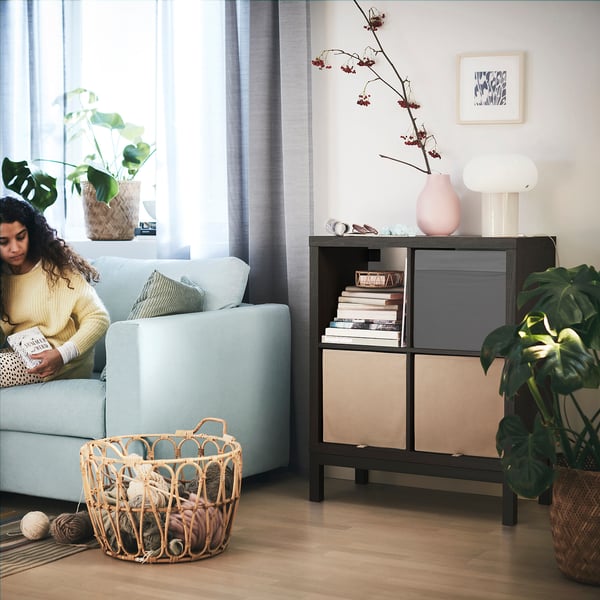 A cosy living room where a person reads on a light blue KALLAX sofa by a large window. Nearby, a KALLAX shelving unit holds books and decor.
