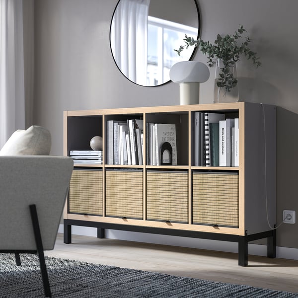 Modern living room with KALLAX shelving unit, light wood & black. Displaying books & decor. Mirror above, grey chair beside, resembling room size.