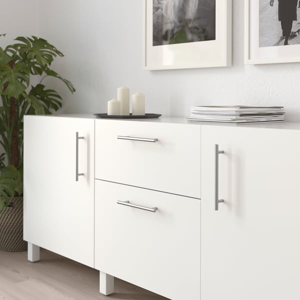 White cabinet with metallic handles, two drawers and two doors. Decorated with candles and books, next to a plant.