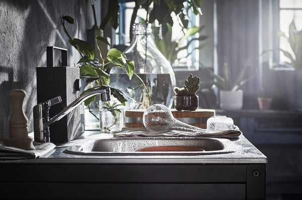Modern kitchen sink with chrome tap, glass containers, and green plants. Sunlight streams through windows.