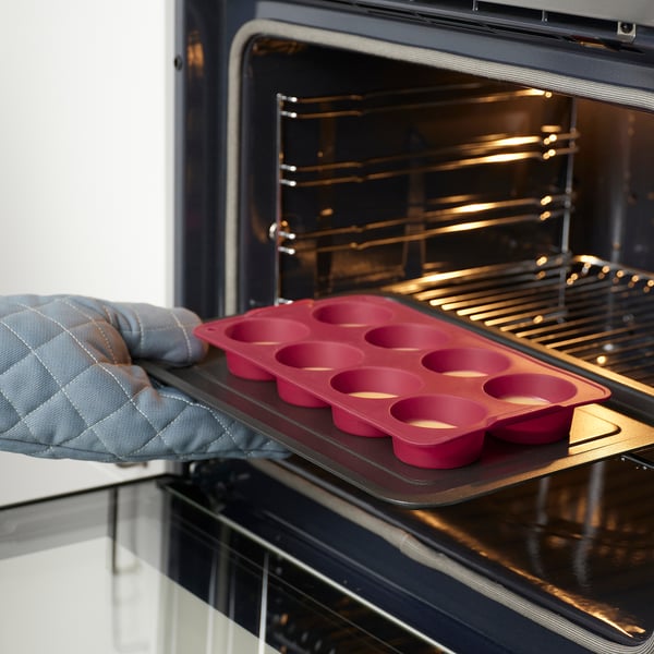 Persons gloved hand placing red silicone muffin tray into oven. Muffin tray holds eight cups for baking.