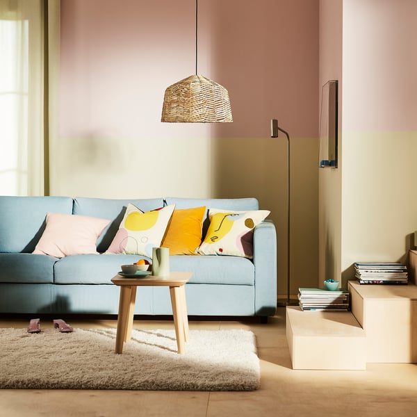 KAPPELAND rattan lamp hangs above blue sofa, casting warm light, pink walls, colourful pillows, white rug.