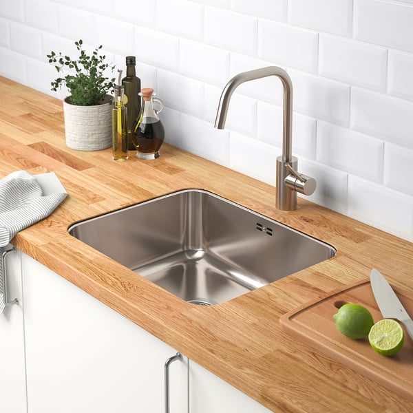 Modern kitchen setup with stainless steel sink, wooden countertop, limes, knife, bottles, and plant. Tap and dish drainer present.