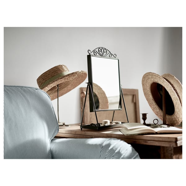 The KARMSUND mirror, black, with intricate top design, sits on a wooden table with a hat display.