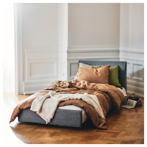 A cosy, compact bed with grey headboard, brown quilt, white blanket, green pillow, and wooden floor; KIVIK.